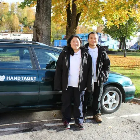 Två medarbetare i Handtagetjackor poserar vid en bil med Handtagets logga